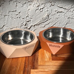 May include: Two pet bowls with stainless steel interiors and geometric, angled exteriors. One bowl is beige, the other is orange. The word "Munnie" is printed on the front of each bowl. The bowls are on a wooden surface.
