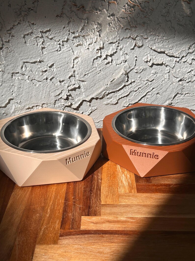 May include: Two pet bowls with stainless steel interiors and geometric, angled exteriors. One bowl is beige, the other is orange. The word "Munnie" is printed on the front of each bowl. The bowls are on a wooden surface.