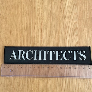 May include: A rectangular black fabric patch featuring the word ARCHITECTS in white capital letters. The patch is positioned above a clear plastic ruler, showing a length of approximately 7.5 inches from the zero mark to the end of the patch.