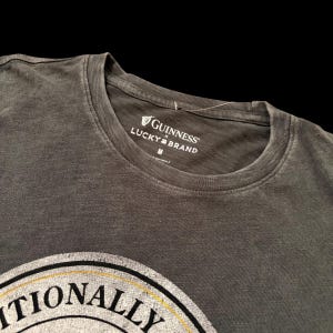 May include: A dark gray t-shirt with a crew neck. The shirt features the Guinness and Lucky Brand logos in white on the collar. The bottom of the shirt displays a partial graphic with the word "ITIONALLY" in black and white.