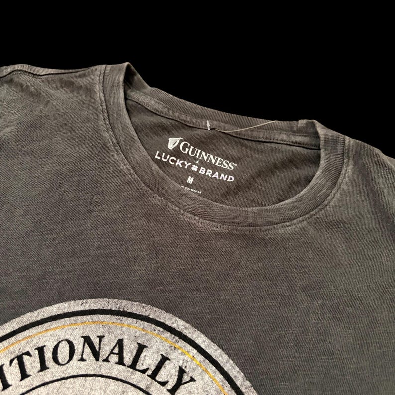 May include: A dark gray t-shirt with a crew neck. The shirt features the Guinness and Lucky Brand logos in white on the collar. The bottom of the shirt displays a partial graphic with the word "ITIONALLY" in black and white.