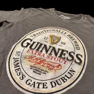 May include: Gray t-shirt featuring a large Guinness Extra Stout label. The label includes the Guinness logo, harp, and text "Traditionally Brewed". The shirt also has the text "St. James's Gate Dublin".