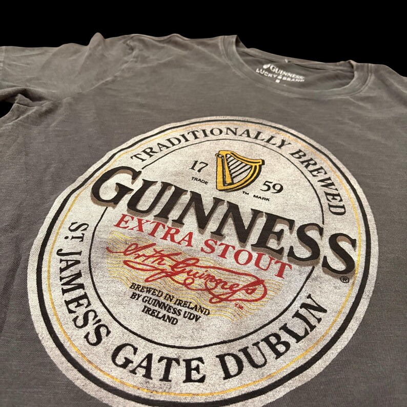 May include: Gray t-shirt featuring a large Guinness Extra Stout label. The label includes the Guinness logo, harp, and text "Traditionally Brewed". The shirt also has the text "St. James's Gate Dublin".