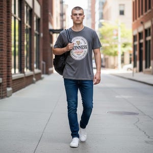 May include: A man wearing a gray t-shirt with a Guinness logo, blue jeans, white sneakers, and a black backpack walks down a city street. The t-shirt features a circular logo with text and a harp symbol. The jeans are a dark wash.