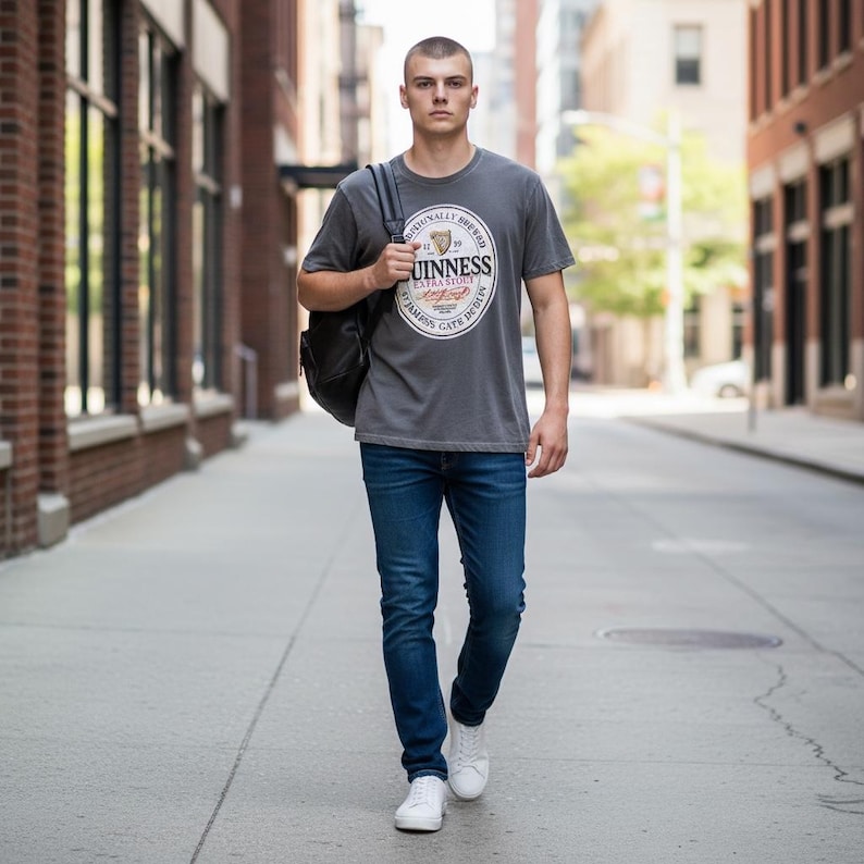 May include: A man wearing a gray t-shirt with a Guinness logo, blue jeans, white sneakers, and a black backpack walks down a city street. The t-shirt features a circular logo with text and a harp symbol. The jeans are a dark wash.