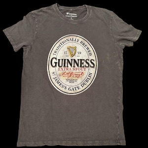 May include: A gray t-shirt featuring the Guinness Extra Stout logo. The logo is a circular design with the Guinness name, a harp, and the text "Traditionally Brewed." The shirt also has the Lucky Brand logo.