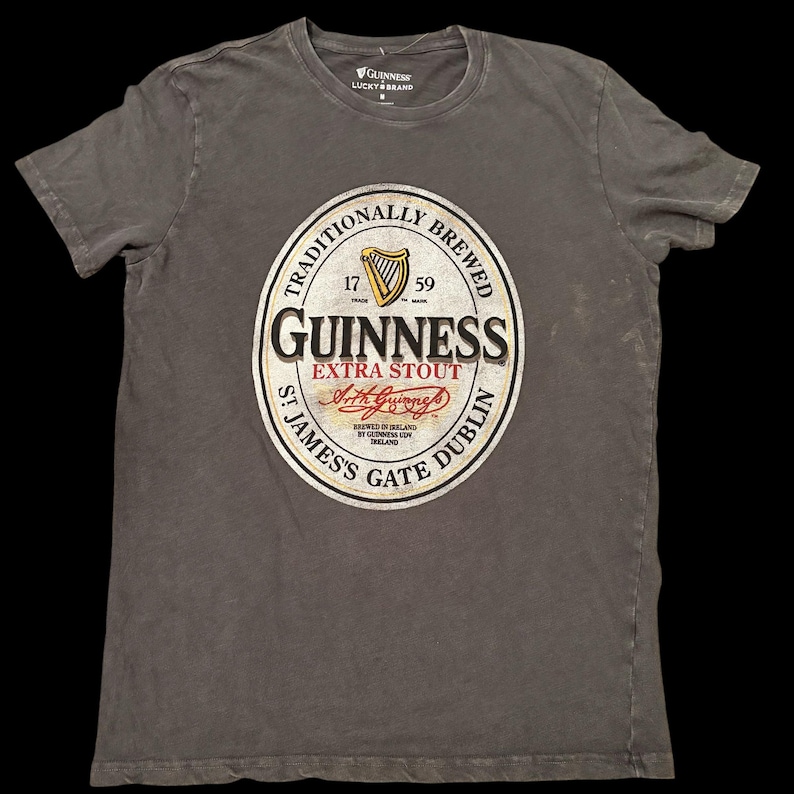 May include: A gray t-shirt featuring the Guinness Extra Stout logo. The logo is a circular design with the Guinness name, a harp, and the text "Traditionally Brewed." The shirt also has the Lucky Brand logo.