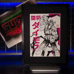 May include: Framed black and white illustration of a robot with Japanese text in pink. The robot is in a dynamic pose, with a raised fist. The artwork is displayed on a silver stand.