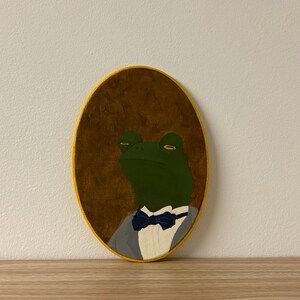 May include: Oval wooden wall art featuring a green frog in a gray suit with a blue bow tie. The frog is painted on a brown background with a yellow border. The artwork is displayed on a wooden surface.