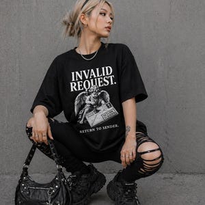 May include: Black t-shirt with the text "INVALID REQUEST." and an angel graphic. The shirt is paired with black ripped jeans, chunky black trainers, and a black handbag. The outfit is completed with a silver necklace and rings.