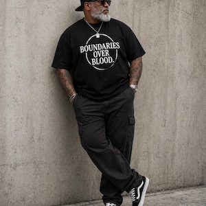May include: Black t-shirt with the text "BOUNDARIES OVER BLOOD" in a white circular design. The person is wearing black cargo pants, black and white sneakers, a black cap, and sunglasses. A silver chain and watch are also visible.