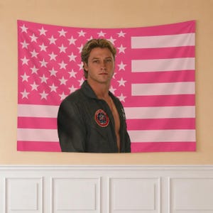 May include: A rectangular wall tapestry featuring a pink and white American flag pattern. Centered on the flag is a portrait of a person with blonde hair wearing a dark green flight jacket with a circular red, white, and blue patch on the chest.