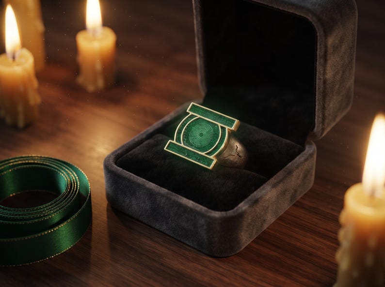 May include: A metallic ring featuring a prominent green rectangular emblem with a circular center design, resting inside a dark velvet-lined jewelry box. The ring has a textured silver-toned band and is displayed alongside green ribbon.