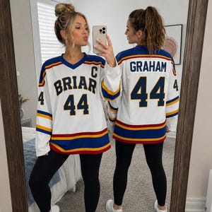 Könnte beinhalten: Ein weißes Eishockeytrikot mit blauen und gelben Streifen an den Ärmeln und am Saum. Die Vorderseite zeigt den Schriftzug BRIAR, die Nummer 44 und ein C. Auf der Rückseite stehen der Name GRAHAM und die Nummer 44 in dunkelblauer Schrift.