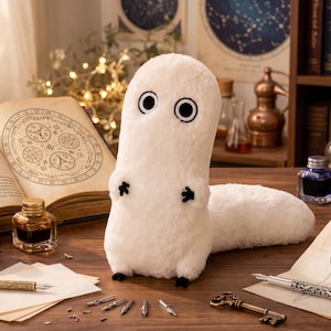 Peut inclure: Une créature en peluche blanche au corps allongé et aux yeux circulaires noirs est posée sur un bureau en bois. Le bureau présente un livre ouvert intitulé Witch Hat Atelier, des encriers en verre bleu, violet et ambre, des plumes et une clé.
