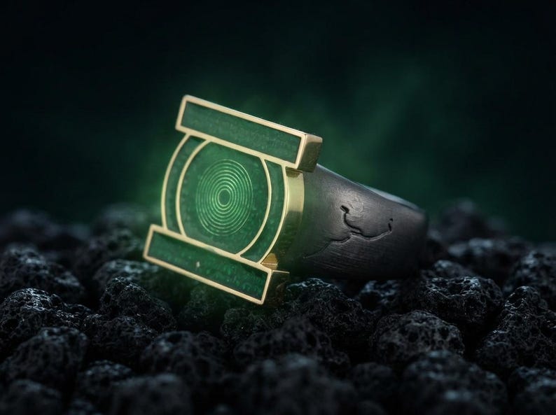 May include: A ring featuring a dark, textured band and a prominent, rectangular green face. The face displays a circular, concentric ring pattern in a lighter green shade, framed by a gold-toned metal border that extends horizontally.