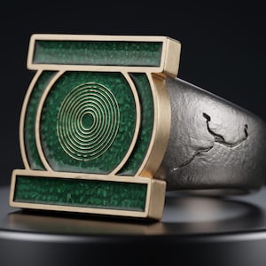 May include: A ring featuring a rectangular gold-toned face with a green textured inlay and a central circular gold spiral design. The band is made of a dark, matte-finished metal with a subtle, irregular etched line pattern along the side.