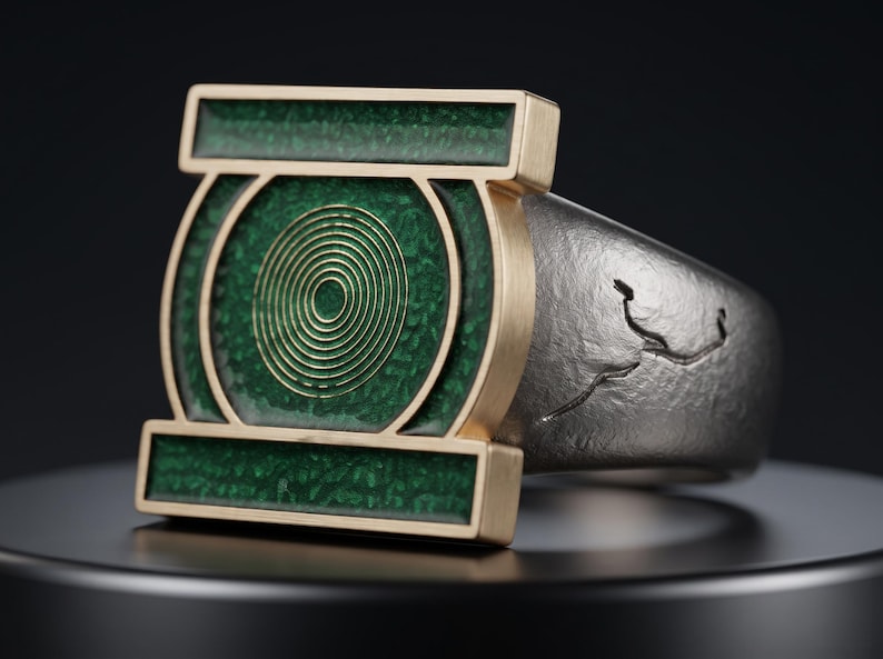 May include: A ring featuring a rectangular gold-toned face with a green textured inlay and a central circular gold spiral design. The band is made of a dark, matte-finished metal with a subtle, irregular etched line pattern along the side.