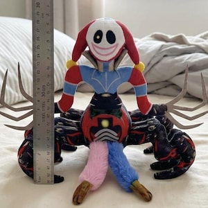 Op de afbeelding: Een knuffel met een wit maskerachtig gezicht met een brede glimlach, rode en blauwe mouwen en een donker, spinnenachtig lichaam met meerdere poten. Een metalen liniaal naast het speelgoed geeft een hoogte van ongeveer 25 centimeter aan.