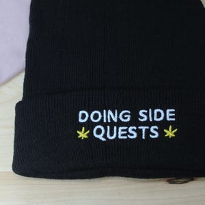 Könnte beinhalten: Eine schwarze Strickmütze mit umgeschlagenem Saum und weißem Sticktext, der DOING SIDE QUESTS lautet, mit einem kleinen gelben sternförmigen Symbol auf jeder Seite des Wortes QUESTS, die eine gerippte Textur im gesamten Stoff aufweist.