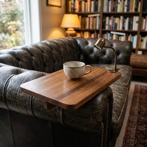 May include: A wooden side table with a cup of coffee on a dark leather couch. A small brass lamp is attached to the side table. The couch is in front of a window and a bookshelf.