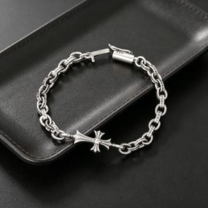 Puede incluir: Una pulsera plateada con un colgante en forma de cruz y un diseño de eslabones de cadena. La pulsera está hecha de un metal brillante y se muestra sobre una superficie negra. El colgante de cruz tiene un pequeño adorno redondo en el centro.
