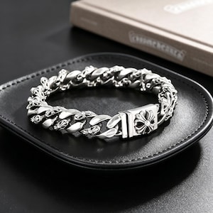 Könnte beinhalten: Ein silberfarbenes Panzerkettenarmband mit verzierten Kreuzdetails. Das Armband wird auf einem schwarzen Ledertablett präsentiert. Der Verschluss hat ein dekoratives Kreuzdesign. Die Glieder des Armbands sind etwa 2 cm breit.