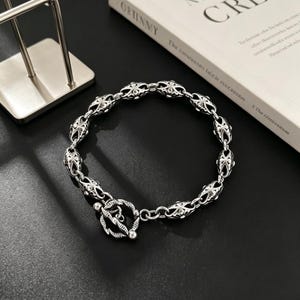 May include: Silver bracelet with a detailed, repeating pattern. The bracelet has a toggle clasp and is shown on a dark surface. The design incorporates floral and cross-like motifs, giving it a textured appearance.