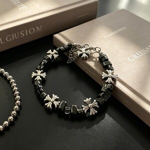 Pode incluir: Uma pulseira de contas pretas com pingentes de cruz prateados. A pulseira é composta por contas pretas quadradas e pingentes decorativos em forma de cruz. Um colar de contas prateadas é visível à esquerda.