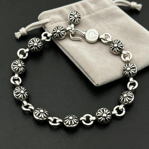 May include: A silver bracelet with daisy-shaped charms. The bracelet is linked together and has a clasp. The bracelet is presented on a grey velvet pouch against a dark background.
