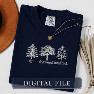 Pode incluir: Camiseta azul marinho com bordados brancos de árvores: abeto, dogwood e tamarack. A camisa tem decote redondo e uma pequena etiqueta. Um colar de ouro com um pingente está sobre a camisa. As palavras "DIGITAL FILE" estão na parte inferior.