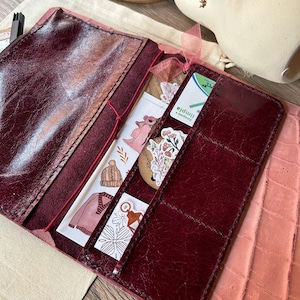 Peut inclure: Journal en cuir bordeaux avec plusieurs poches et fentes pour cartes. Le journal ouvert affiche des autocollants décoratifs, des cartes et un ruban rose. Le journal est sur une surface rose texturée.