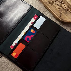 Può includere: Un portafoglio in pelle nera con uno scomparto con cerniera, diversi scomparti per carte e una tasca verticale. Il portafoglio è aperto e mostra un inserto di carta a quadretti, una carta rossa, una carta di pagamento blu e rossa.