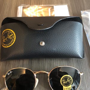 May include: Round Ray-Ban sunglasses with gold frames and dark lenses, displayed with a black leather case and cleaning cloth. The sunglasses have the Ray-Ban logo on the lens and frame.