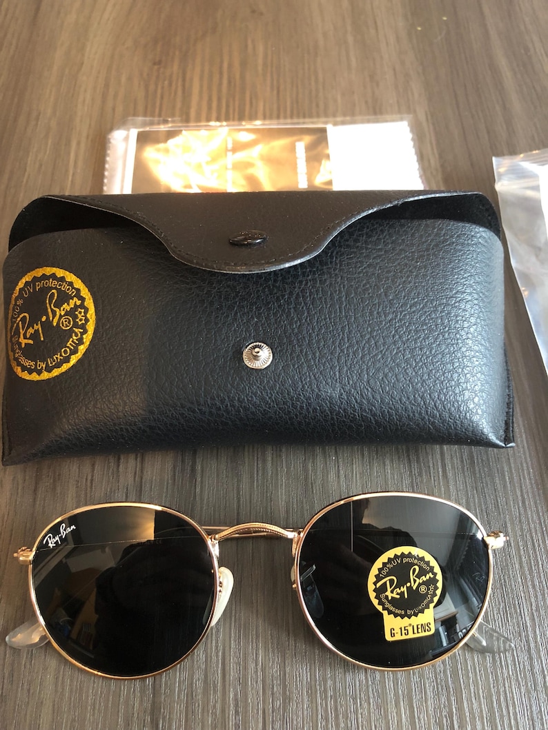 May include: Round Ray-Ban sunglasses with gold frames and dark lenses, displayed with a black leather case and cleaning cloth. The sunglasses have the Ray-Ban logo on the lens and frame.