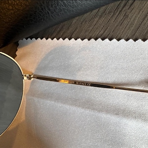 May include: Close-up of gold-toned sunglasses with round lenses. The frame is thin and delicate, with the text "MADE IN ITALY CE" visible on the temple. The sunglasses are resting on a light-colored, textured surface.