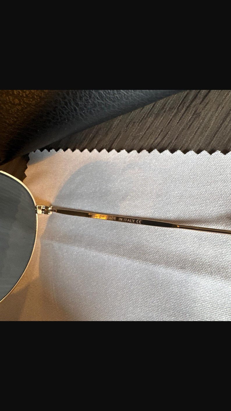 May include: Close-up of gold-toned sunglasses with round lenses. The frame is thin and delicate, with the text "MADE IN ITALY CE" visible on the temple. The sunglasses are resting on a light-colored, textured surface.