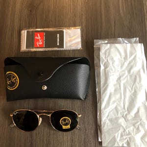 May include: A set of Ray-Ban sunglasses with a black leather case, cleaning cloth, and packaging. The sunglasses have gold frames and dark lenses. The case has a gold logo. The cleaning cloth has the Ray-Ban logo and website address.