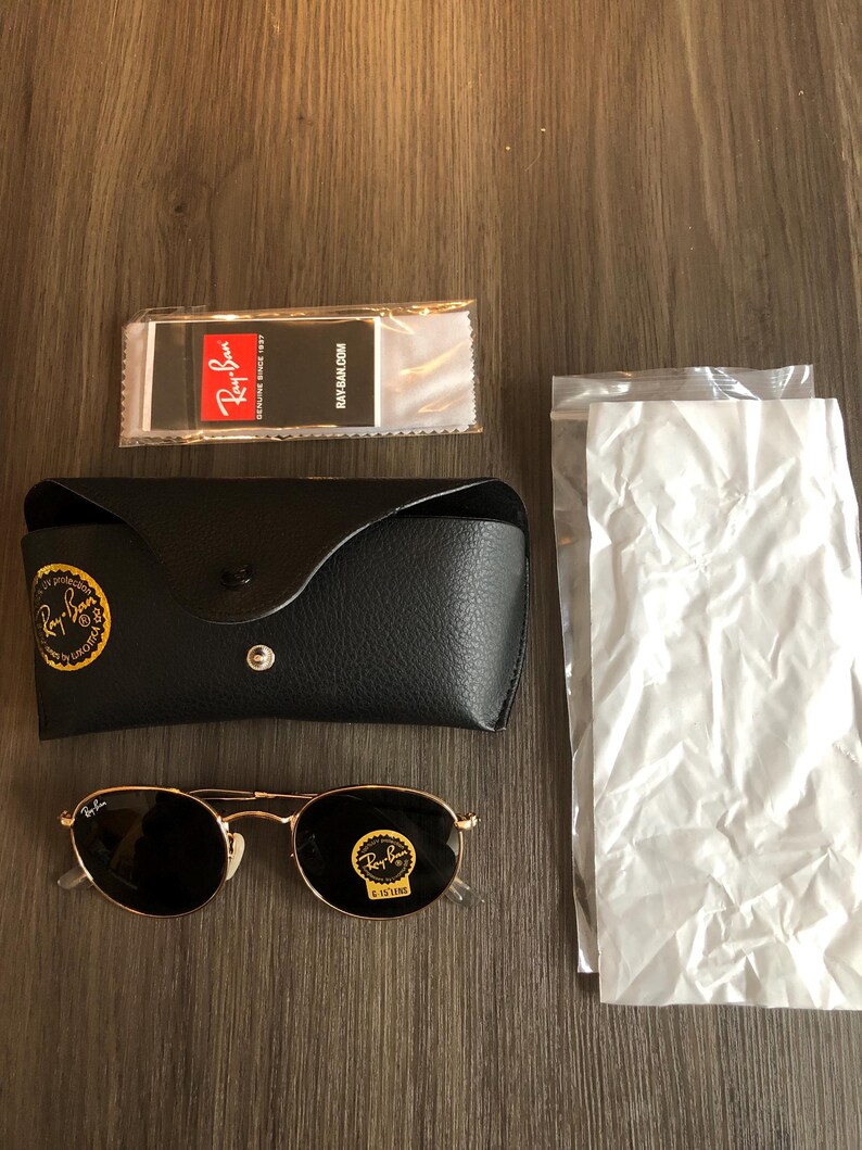 May include: A set of Ray-Ban sunglasses with a black leather case, cleaning cloth, and packaging. The sunglasses have gold frames and dark lenses. The case has a gold logo. The cleaning cloth has the Ray-Ban logo and website address.