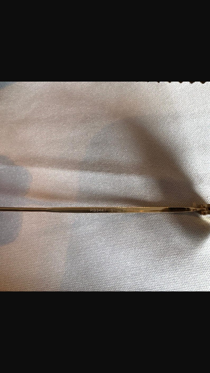 May include: Close-up of a gold-colored eyeglass arm. The text "RB3447 51" is printed on the arm. The background is a light-colored, textured fabric.
