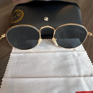 May include: Gold-rimmed sunglasses with dark lenses, a black leather case, and a cleaning cloth. The sunglasses have a classic oval shape and a bridge over the nose. The case has a snap closure and a gold logo.