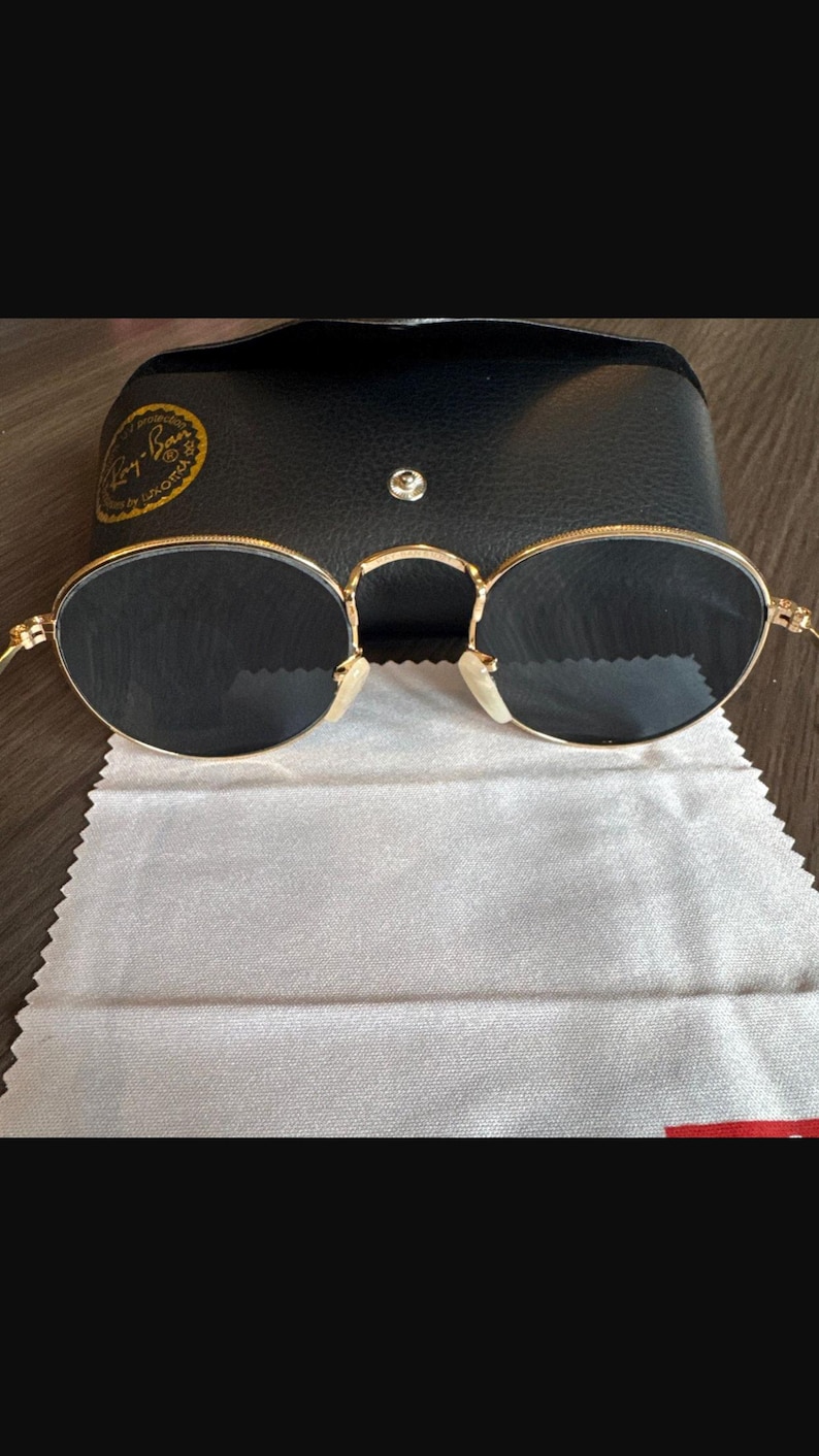 May include: Gold-rimmed sunglasses with dark lenses, a black leather case, and a cleaning cloth. The sunglasses have a classic oval shape and a bridge over the nose. The case has a snap closure and a gold logo.
