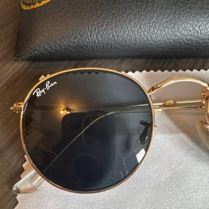 May include: Gold-rimmed sunglasses with dark lenses, resting on a gray cloth. The Ray-Ban logo is visible on the lens. A black leather case is in the background. The sunglasses have a classic, round shape.