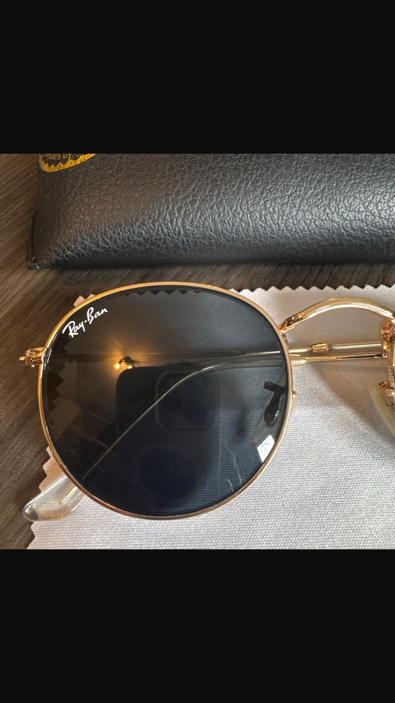 May include: Gold-rimmed sunglasses with dark lenses, resting on a gray cloth. The Ray-Ban logo is visible on the lens. A black leather case is in the background. The sunglasses have a classic, round shape.