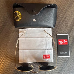 May include: Gold-framed sunglasses with black lenses, a black leather case, a cleaning cloth, and a product information card. The sunglasses and case feature the Ray-Ban logo. The case is closed with a snap.