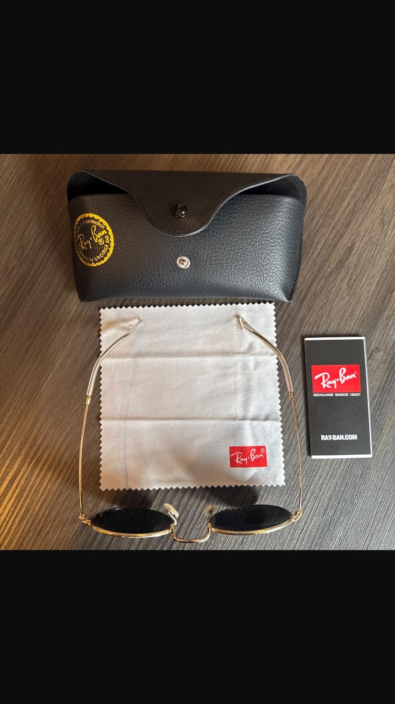 May include: Gold-framed sunglasses with black lenses, a black leather case, a cleaning cloth, and a product information card. The sunglasses and case feature the Ray-Ban logo. The case is closed with a snap.