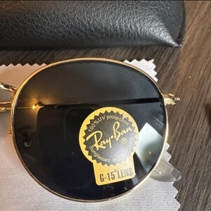 May include: Gold-rimmed Ray-Ban sunglasses with dark lenses. The sunglasses have a round shape and a sticker that reads "100% UV protection Ray-Ban Sunglasses G-15 LENS". The sunglasses are on a white cloth.