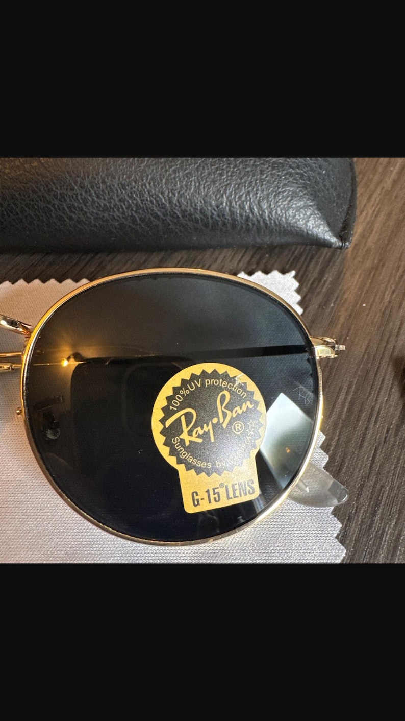 May include: Gold-rimmed Ray-Ban sunglasses with dark lenses. The sunglasses have a round shape and a sticker that reads "100% UV protection Ray-Ban Sunglasses G-15 LENS". The sunglasses are on a white cloth.