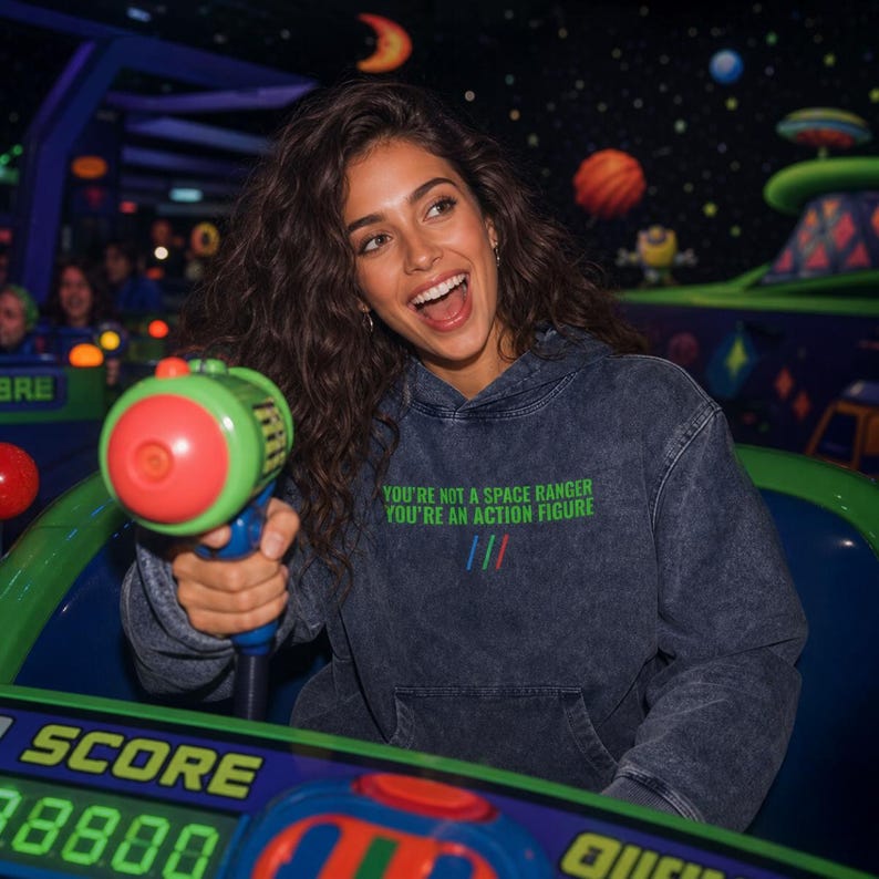 K&ouml;nnte beinhalten: Ein dunkelgrauer, s&auml;uregewaschener Kapuzenpullover mit dem gr&uuml;nen gestickten Text "YOU'RE NOT A SPACE RANGER YOU'RE AN ACTION FIGURE" auf der Brust, begleitet von drei kleinen vertikalen Linien in Blau, Rot und Gr&uuml;n unter dem Text.
