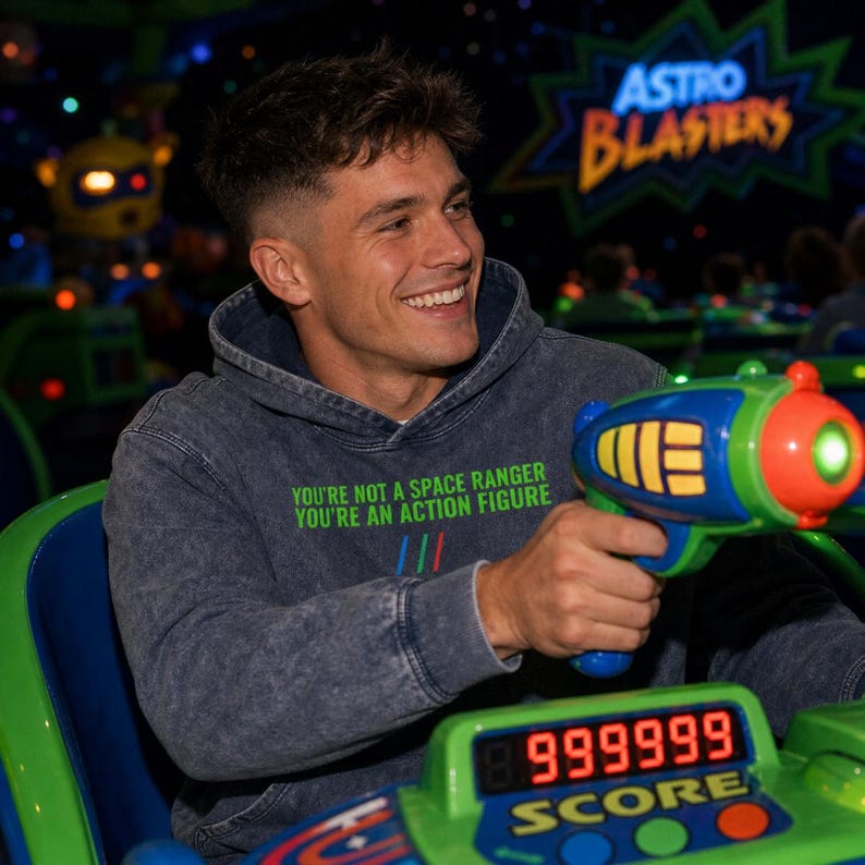 K&ouml;nnte beinhalten: Ein dunkelgrauer, s&auml;uregewaschener Kapuzenpullover mit dem gr&uuml;nen Text "YOU'RE NOT A SPACE RANGER YOU'RE AN ACTION FIGURE" und drei kleinen vertikalen Linien in Blau, Rot und Gr&uuml;n, getragen beim Halten eines blauen, gelben und gr&uuml;nen Spielzeug-Blasters.